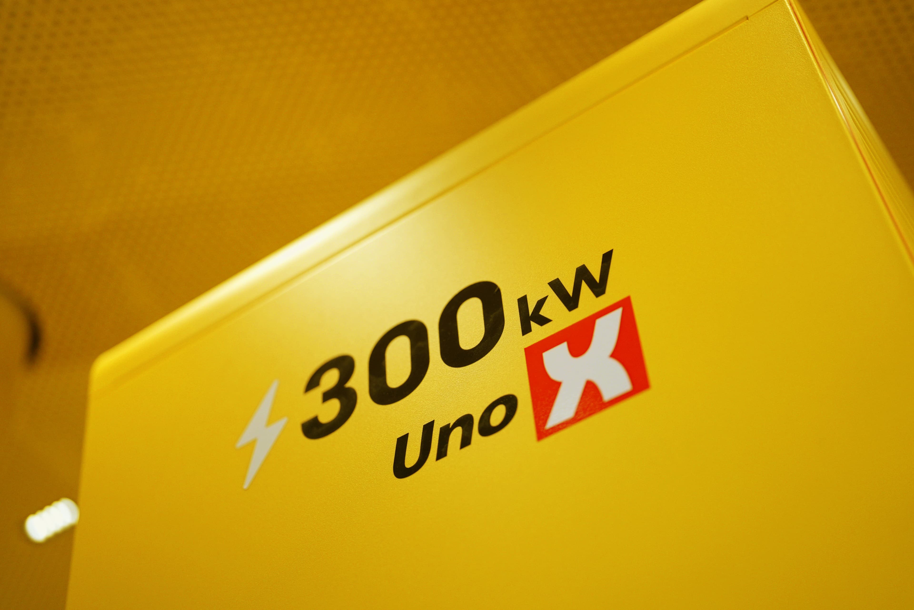 En gul ladestationsenhed med teksten "300 kW Uno X" trykt på forsiden. Teksten inkluderer et lynsymbol, der indikerer hurtig opladning, og et rødt og hvidt Uno X-logo.